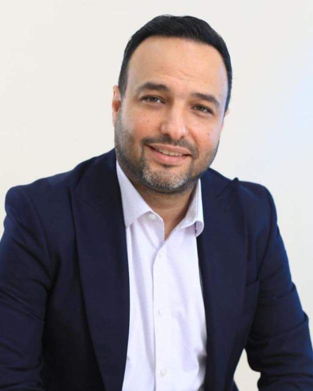 Firas Bou Diab, a man with short dark hair and a beard, smiles at the camera in his navy blazer and white shirt against a plain background.