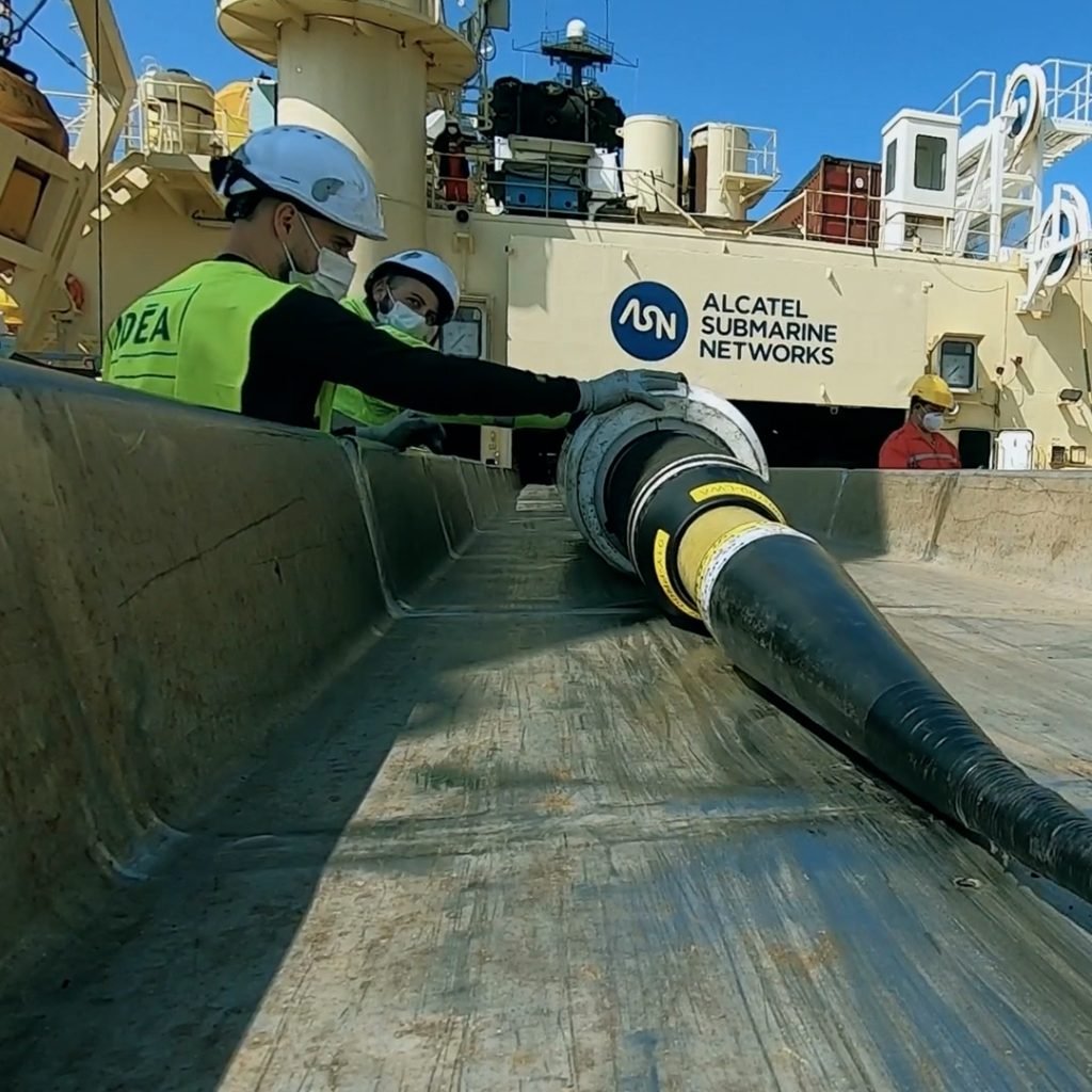 Workers from Alcatel Submarine Networks expertly manage a large underwater cable on the ship's deck, donning safety gear and helmets to ensure precision and safety in their critical maritime task.