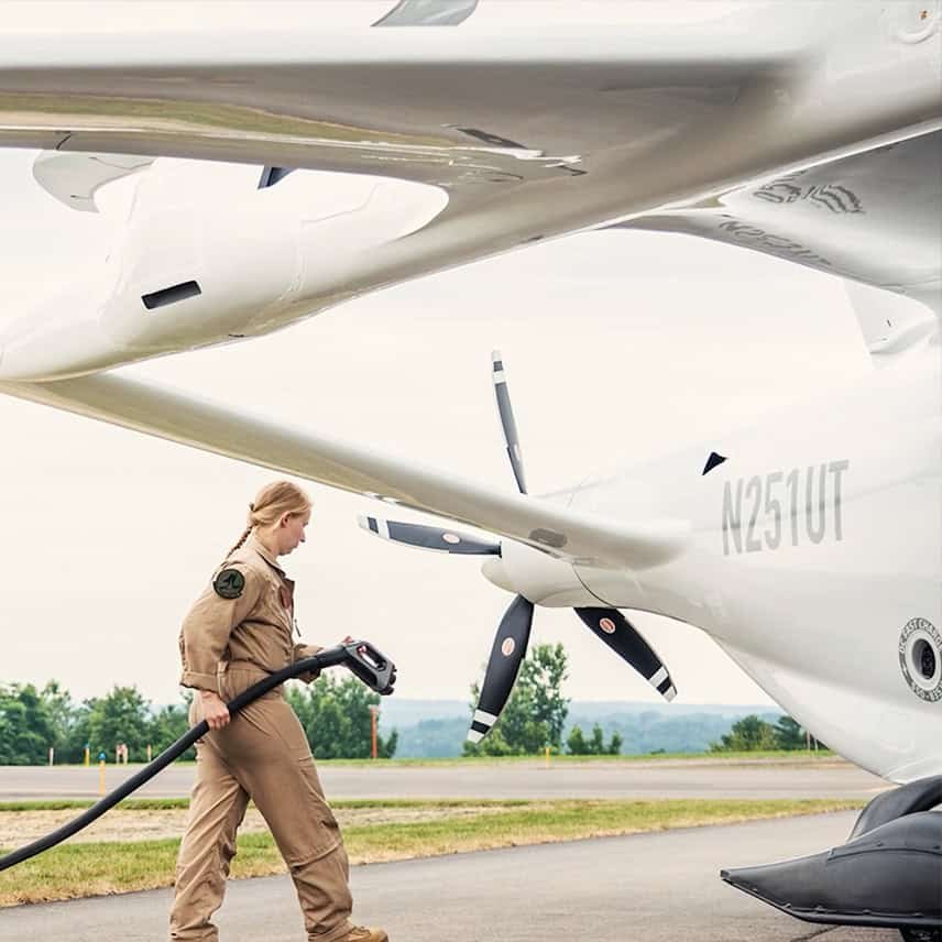 A person in a brown jumpsuit is refueling a white aircraft with large propellers, bearing registration number N251UT on the Albany International tarmac. Trees line the background under a cloudy sky, setting the scene for this busy airport moment.