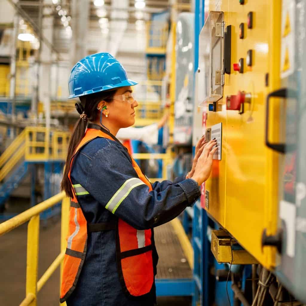 A person in a blue hard hat and orange safety vest operates machinery in an industrial setting.