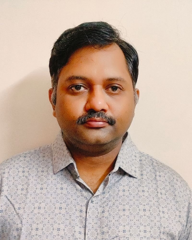 Prashant Atmakuri, with short dark hair and a mustache, stands confidently in a patterned shirt against a plain off-white background.