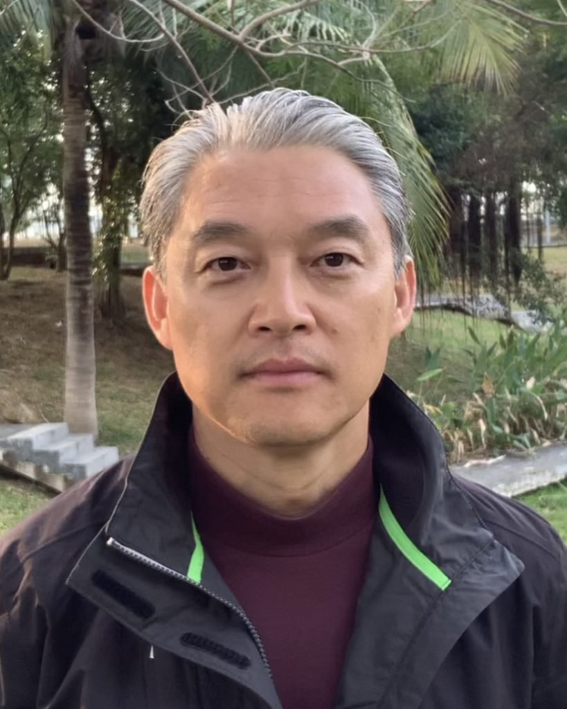 Dr. Lihui Xu, a distinguished gentleman with gray hair, stands outdoors in a black jacket, surrounded by verdant trees and lush grass.