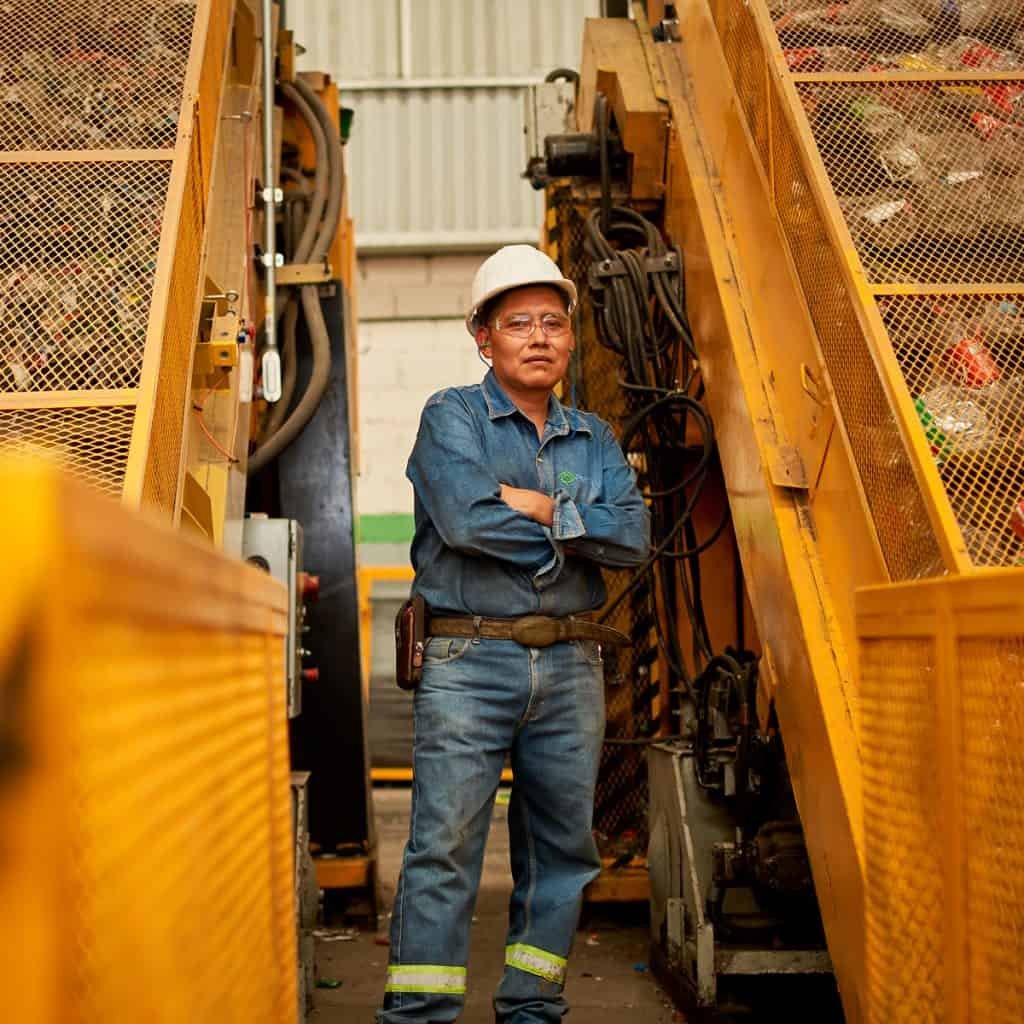 A person in a denim outfit and white hard hat stands between industrial machinery, arms crossed.