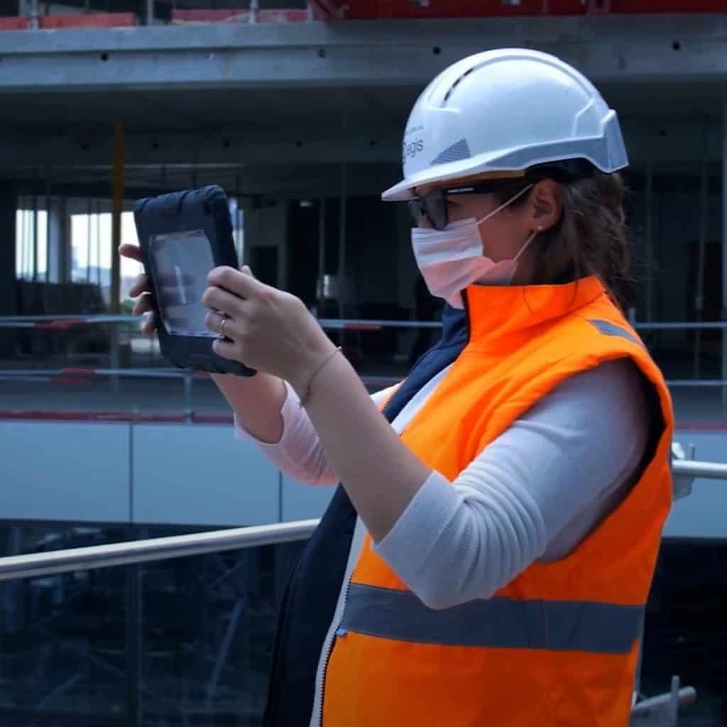 A person in an orange safety vest and white helmet uses a tablet at a construction site.