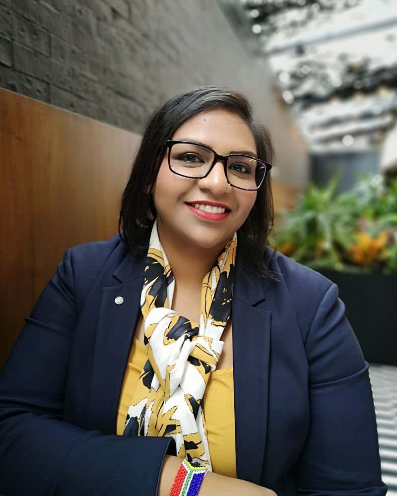 Jeshika Moonsamy, wearing glasses and a dark blazer with a patterned scarf, smiles while sitting indoors surrounded by lush plants.