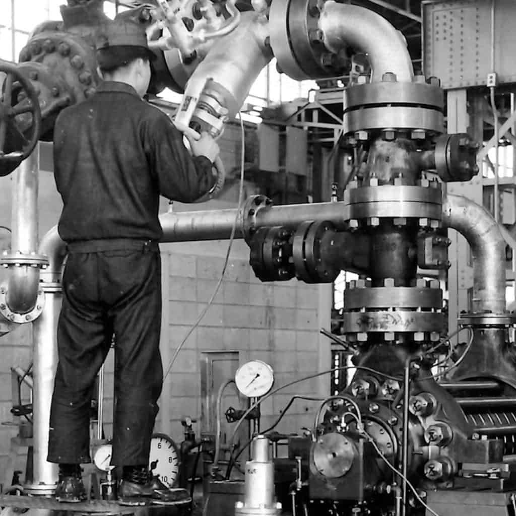 Worker in uniform checks a large industrial valve system inside a factory, surrounded by pipes and gauges.