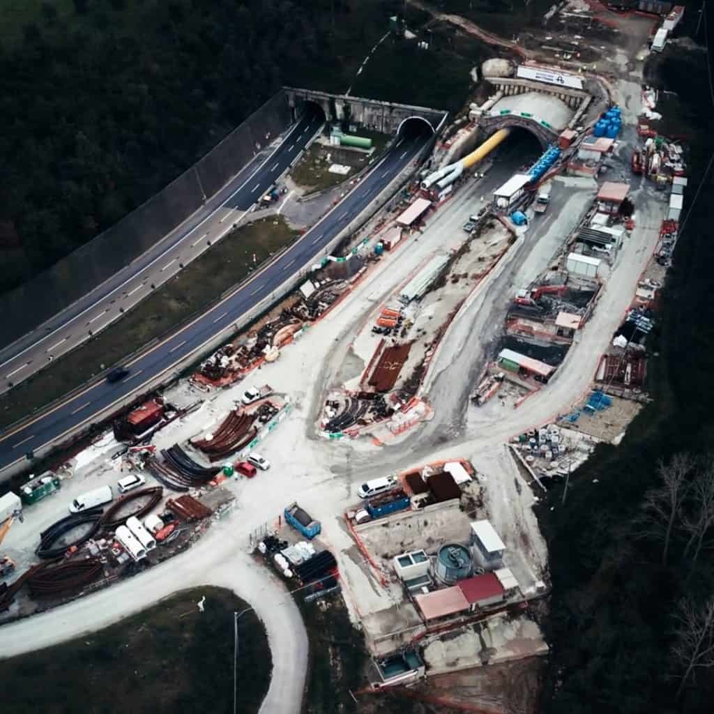 Aerial view of an Autostrade per I’Italia highway construction site featuring tunnels, various equipment, and materials scattered among the lush greenery.