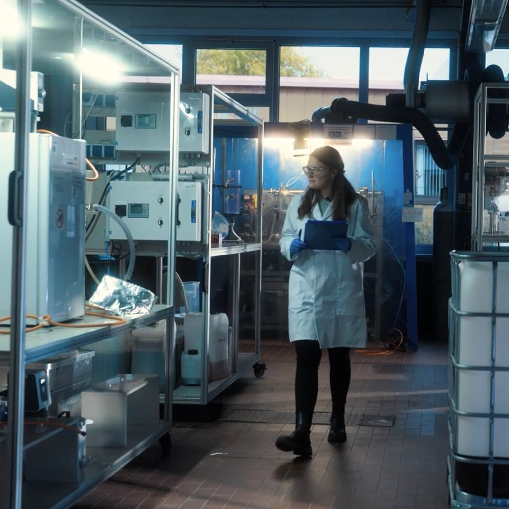 A person in a lab coat and gloves examines equipment in a laboratory filled with machines and bright lighting.