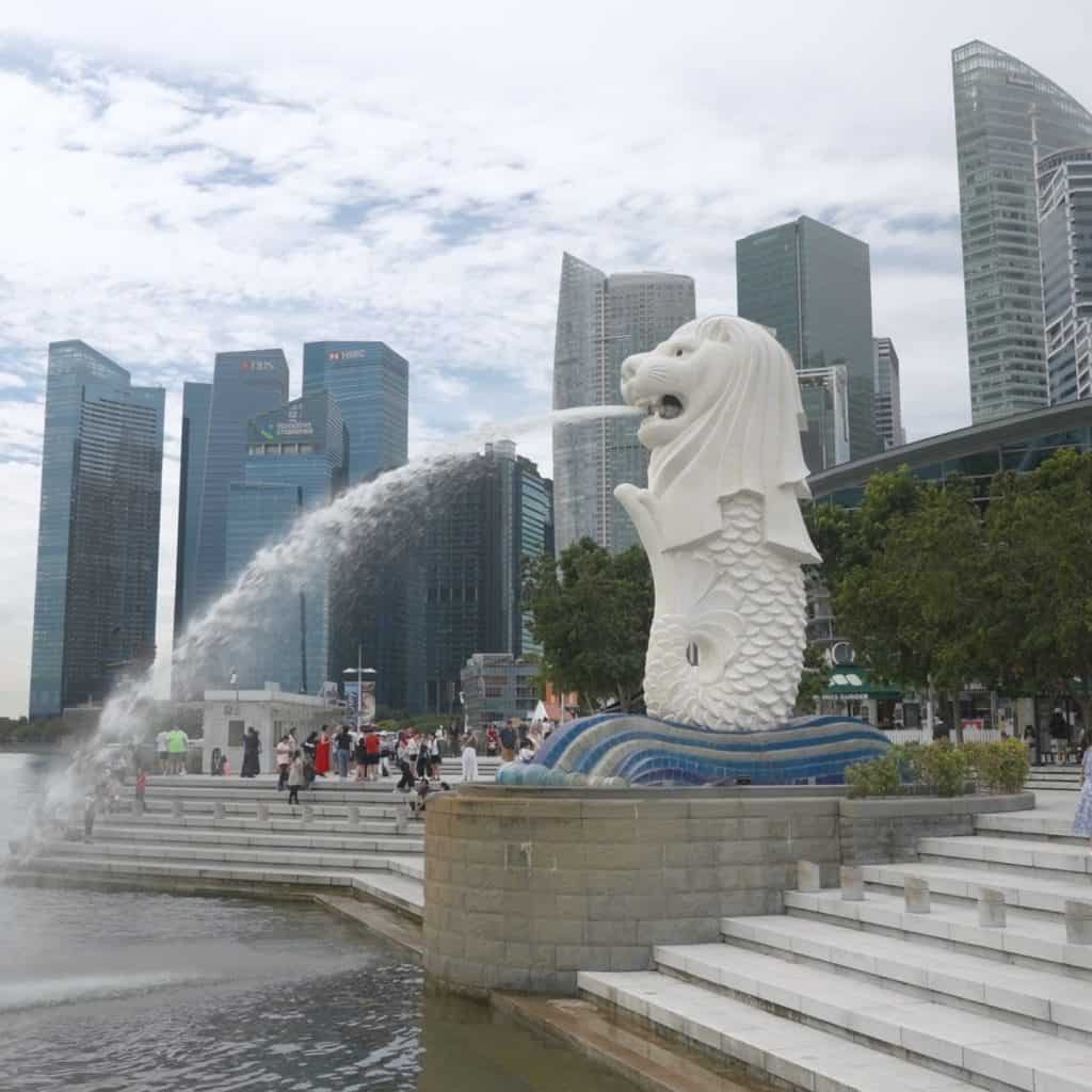Merlion statue spouts water in Marina Bay, Singapore, surrounded by skyscrapers and tourists. Steps and greenery are in the foreground.