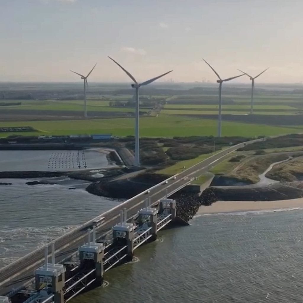 Aerial view of wind turbines on a coastline near a dam. Roads and a river are visible, with lush green fields in the background under a clear sky.