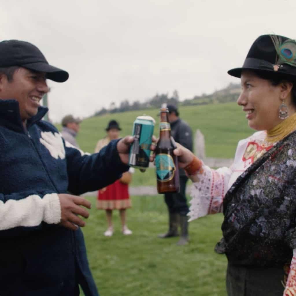 Two people outdoors clink a can and a bottle, wrapped in warm clothing. A grassy field and other revelers enjoying the day in Ecuador are visible in the background, perhaps savoring local flavors from Cervecería Nacional.