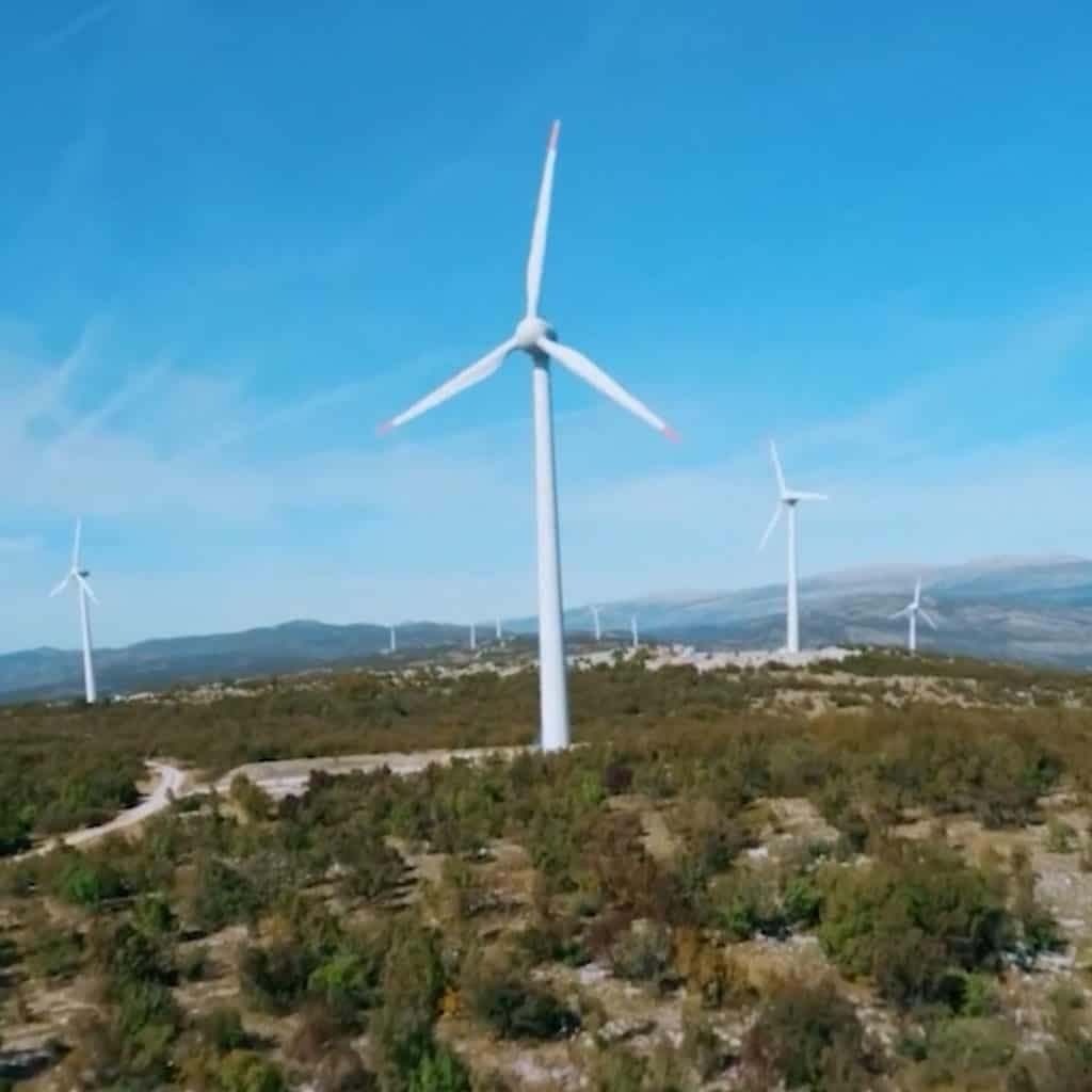 Wind turbines on a hilly landscape with green vegetation. Blue sky above, with scattered clouds.