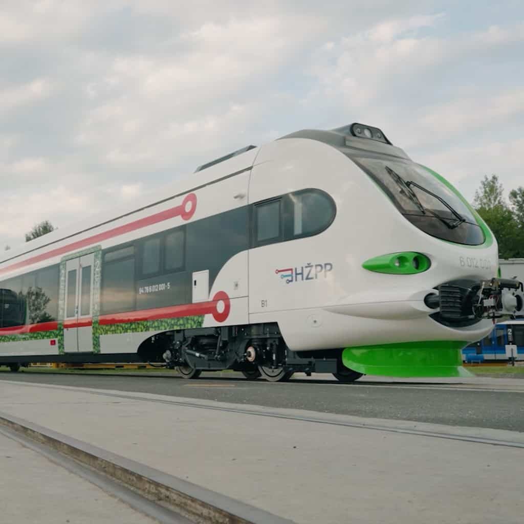 Modern train parked on railway tracks in an outdoor setting, featuring a sleek design with white, black, red, and green accents. Sky and trees are visible in the background.