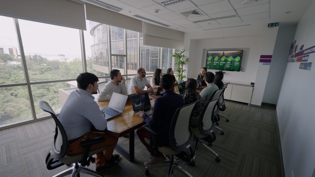 People in a conference room have a meeting. Laptops and papers are on the table, and a presentation is displayed on a wall screen. Large windows show a view of trees and a building outside.