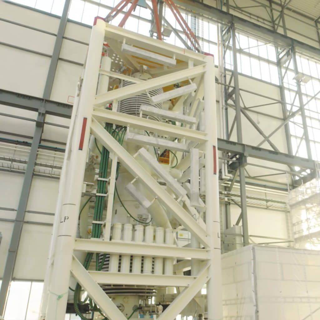 Industrial machinery with a lattice framework suspended indoors, featuring various components, pipes, and connectors against a backdrop of large windows and steel beams.