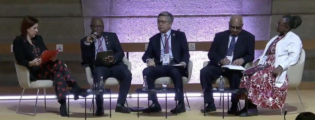 Five people are seated on a stage in a panel discussion, one speaking into a microphone. Dressed in formal attire, they expertly engage in the vibrant exchange of ideas. Broadcast live as a webcast, their conversation offers insights shaping the landscape of 2025.