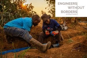 Two engineers in work clothes install pipes in a trench, surrounded by tools and lush vegetation. The Engineers Without Borders logo is proudly displayed in the top right corner, highlighting the international scope of their impactful work.