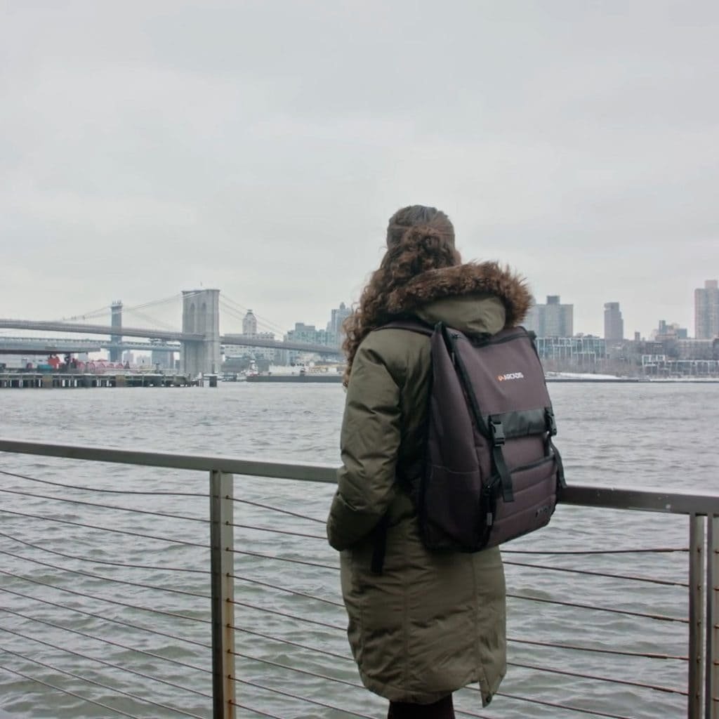 A person wearing a green coat and backpack stands by a railing, overlooking a river with bridges and city buildings in the background.
