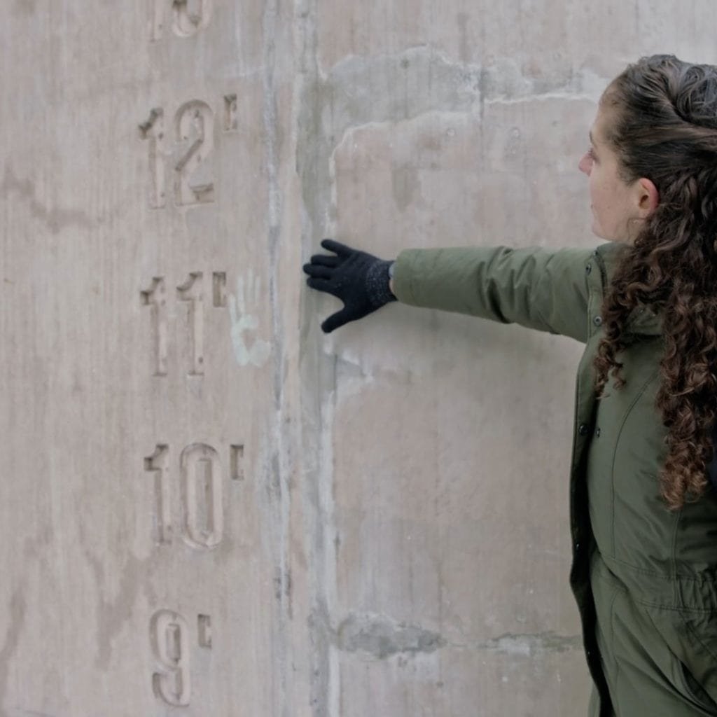 A person wearing a green coat and black gloves stands beside a concrete wall with height markings in feet from 9 to 13. They are pointing at the 11-foot mark.